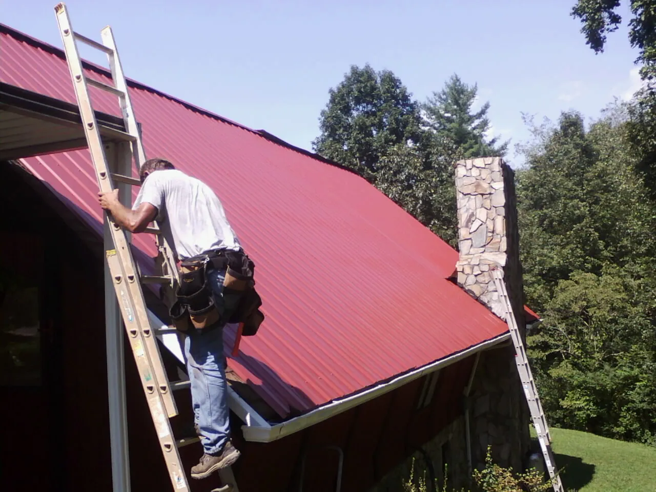 Roofer on a ladder beside a red metal roof and stone chimney — Soffit & Fascia Repair by Roofing Brothers in Charles City